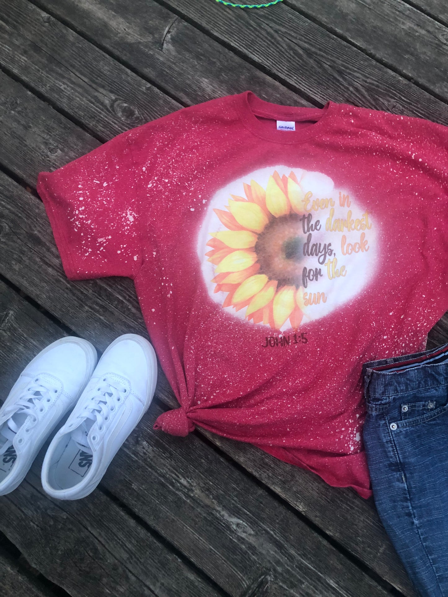 Red T-shirt with bleach splatter. Featuring a half sunflower and script to match for the other half in color and shape. It reads " Even in the darkest of days, look for the sun. " Under the sunflower it reads John 1:5